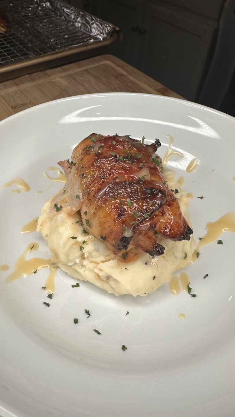 A Bacon Wrapped Honey Mustard Chicken breast with a glossy glaze sits atop creamy mashed potatoes, garnished with chopped herbs and drizzled sauce, served on a white plate.