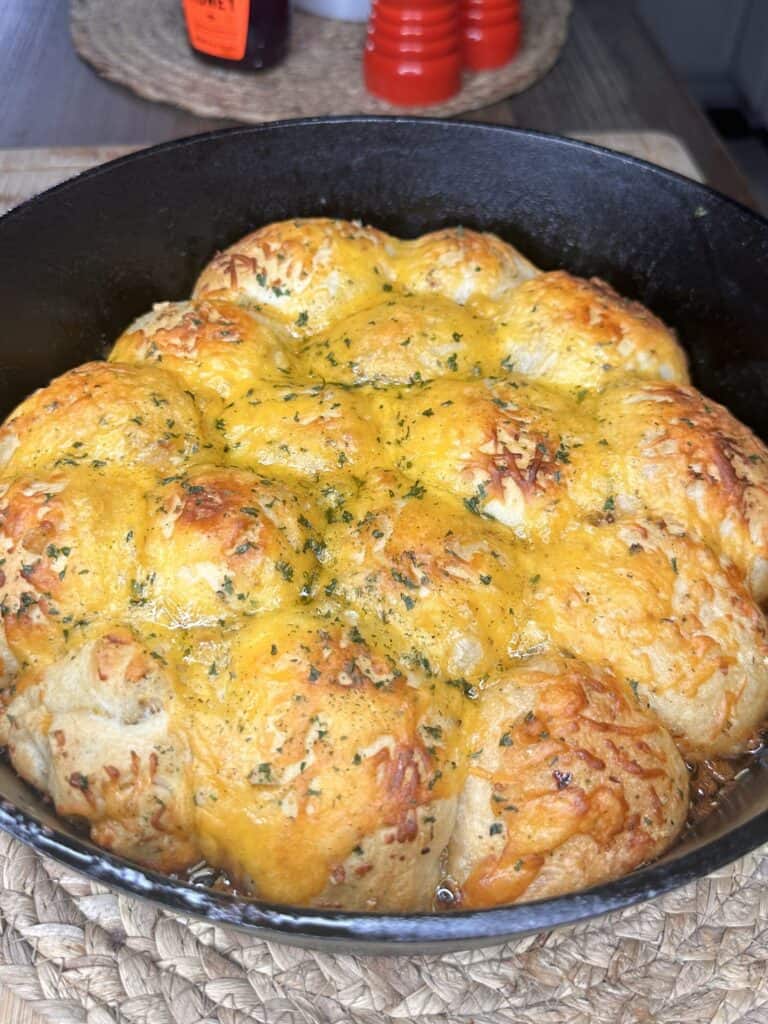 A cast iron skillet filled with Easy Cheesy Garlic Bombs—golden, cheesy garlic bread rolls topped with melted cheese and herbs—rests on a woven placemat.