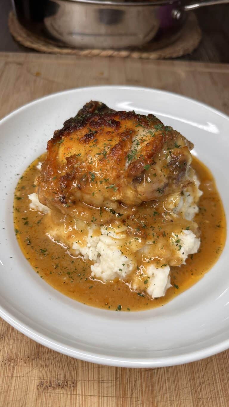 A plate of mashed potatoes topped with a browned, saucy Smothered Chicken thigh and garnished with herbs, with extra orange-brown gravy pooling around the base. A pot is visible in the background on a wooden surface.