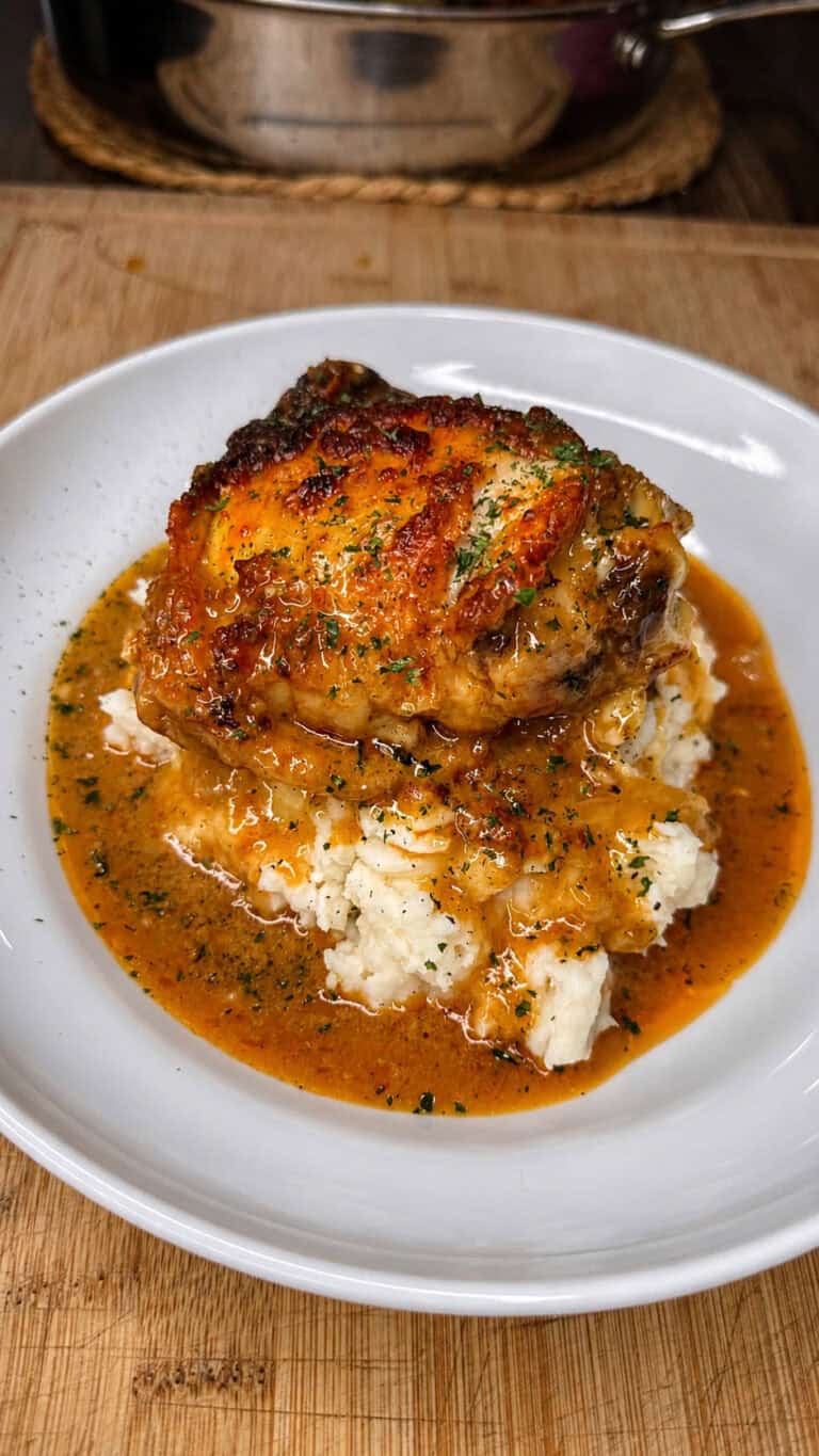 A piece of crispy Smothered Chicken sits atop mashed potatoes, topped with rich brown gravy and herbs, served on a white plate on a wooden table.