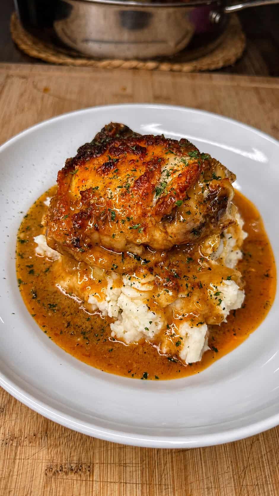 A piece of crispy Smothered Chicken sits atop mashed potatoes, topped with rich brown gravy and herbs, served on a white plate on a wooden table.