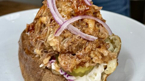 A Pigs in a Potato special: a baked potato loaded with pulled barbecue meat, coleslaw, pickle slices, and topped with thinly sliced red onions, served on a white plate.