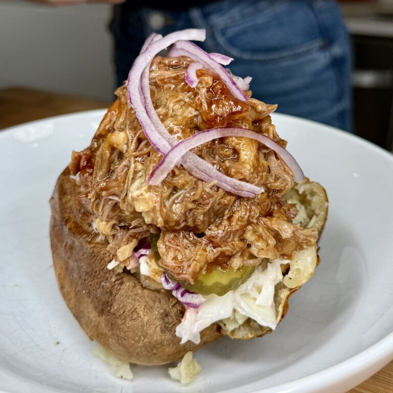 A Pigs in a Potato special: a baked potato loaded with pulled barbecue meat, coleslaw, pickle slices, and topped with thinly sliced red onions, served on a white plate.