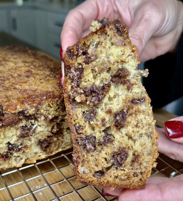 A close-up of hands holding a thick slice of moist Melissa Jo's Banana Bread filled with chocolate chips. The rest of the loaf sits on a cooling rack in a kitchen in the background.