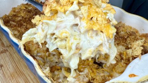 A serving spoon lifts a cheesy noodle casserole, The Best Tuna Casserole, topped with crispy crumbs from a ceramic baking dish on a wooden surface.