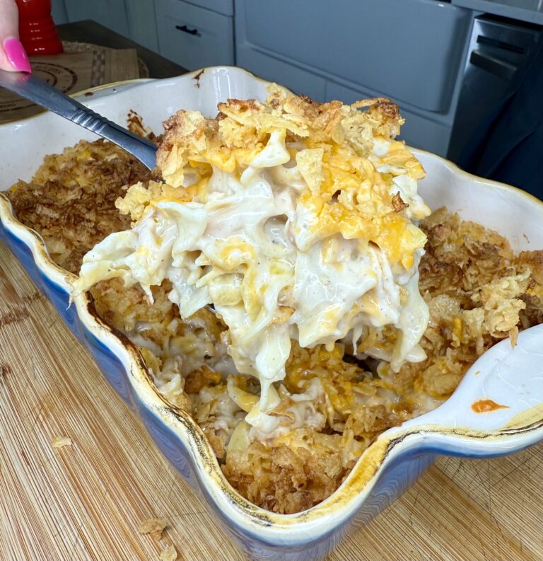 A serving spoon lifts a cheesy noodle casserole, The Best Tuna Casserole, topped with crispy crumbs from a ceramic baking dish on a wooden surface.