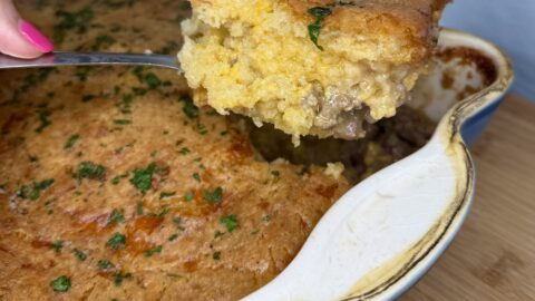 A close-up of a casserole dish filled with a baked cornbread-topped meat pie, reminiscent of a Cheeseburger on a Biscuit Casserole. A spoon lifts a serving, revealing the golden crust, ground meat filling, and sprinkled herbs on top.