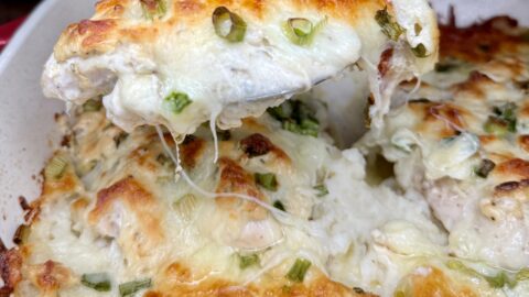 A close-up of a cheesy One Pan Chicken and Mash casserole being served from a dish, showing melted cheese, green onions, and a golden-brown, bubbly top layer.