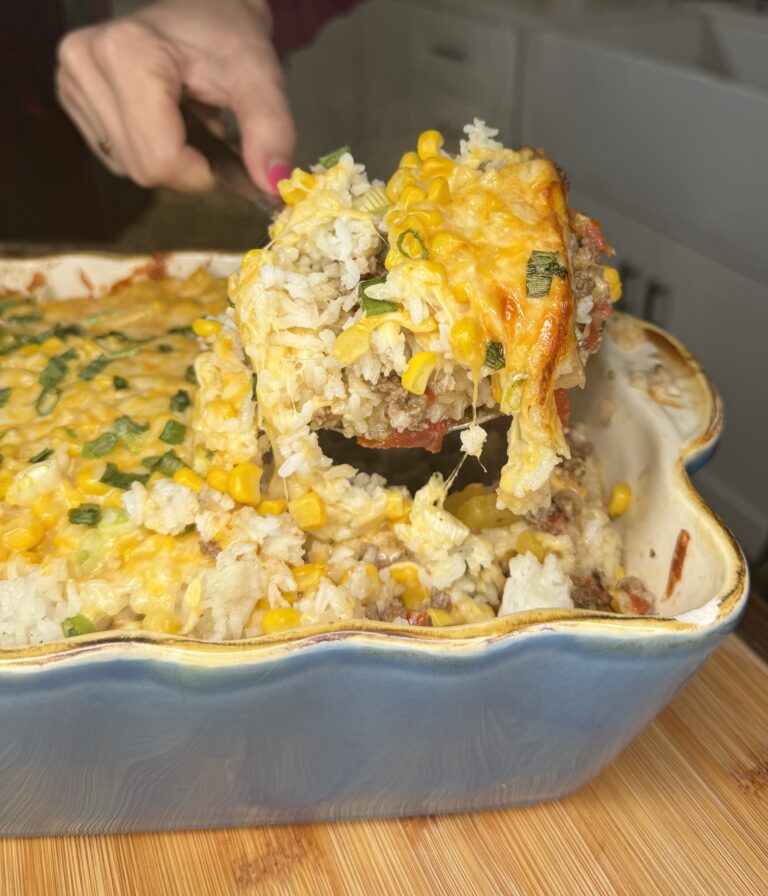 A hand serves a cheesy Texas Rice Casserole with corn, ground meat, and green onions from a blue baking dish on a wooden surface. The casserole is topped with melted cheese and looks freshly baked.