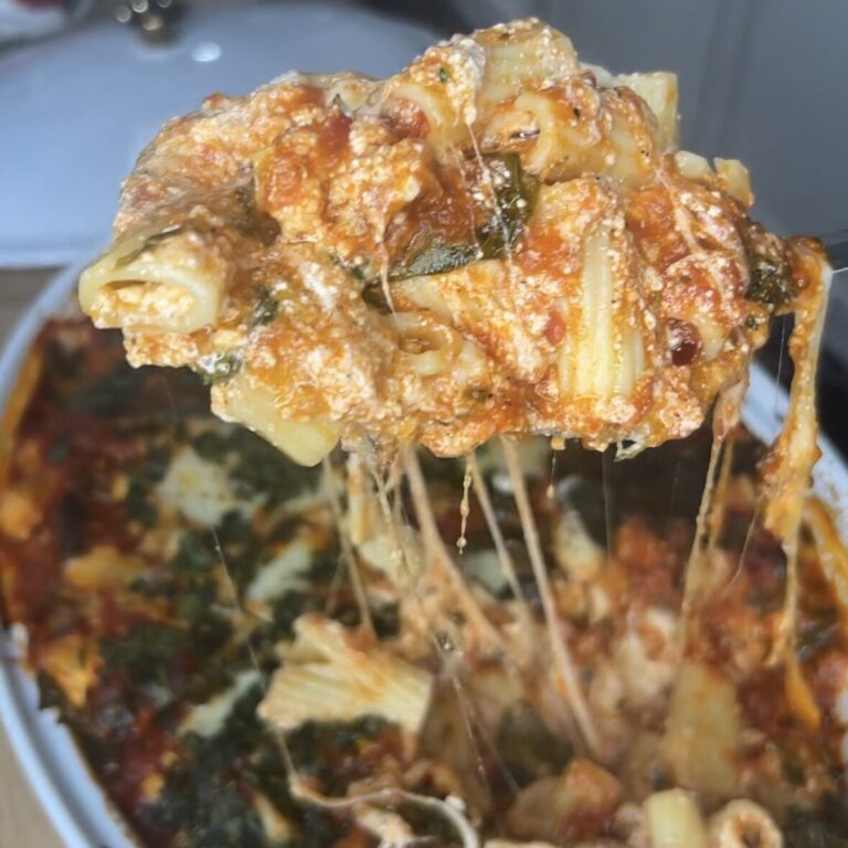 A close-up of Melissa Jo's Baked Ziti being lifted with a spoon, showing gooey, melted cheese stretching from the dish below. The pasta is coated in tomato sauce with visible herbs and creamy ricotta.