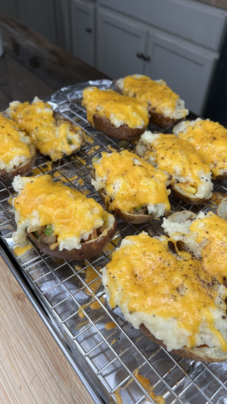 A tray of Shepard's Pie Twice Baked Potatoes with melted cheddar cheese and seasoning rests on a wire rack in a kitchen. The potato halves are stuffed and slightly overflowing with a creamy, flavorful filling.