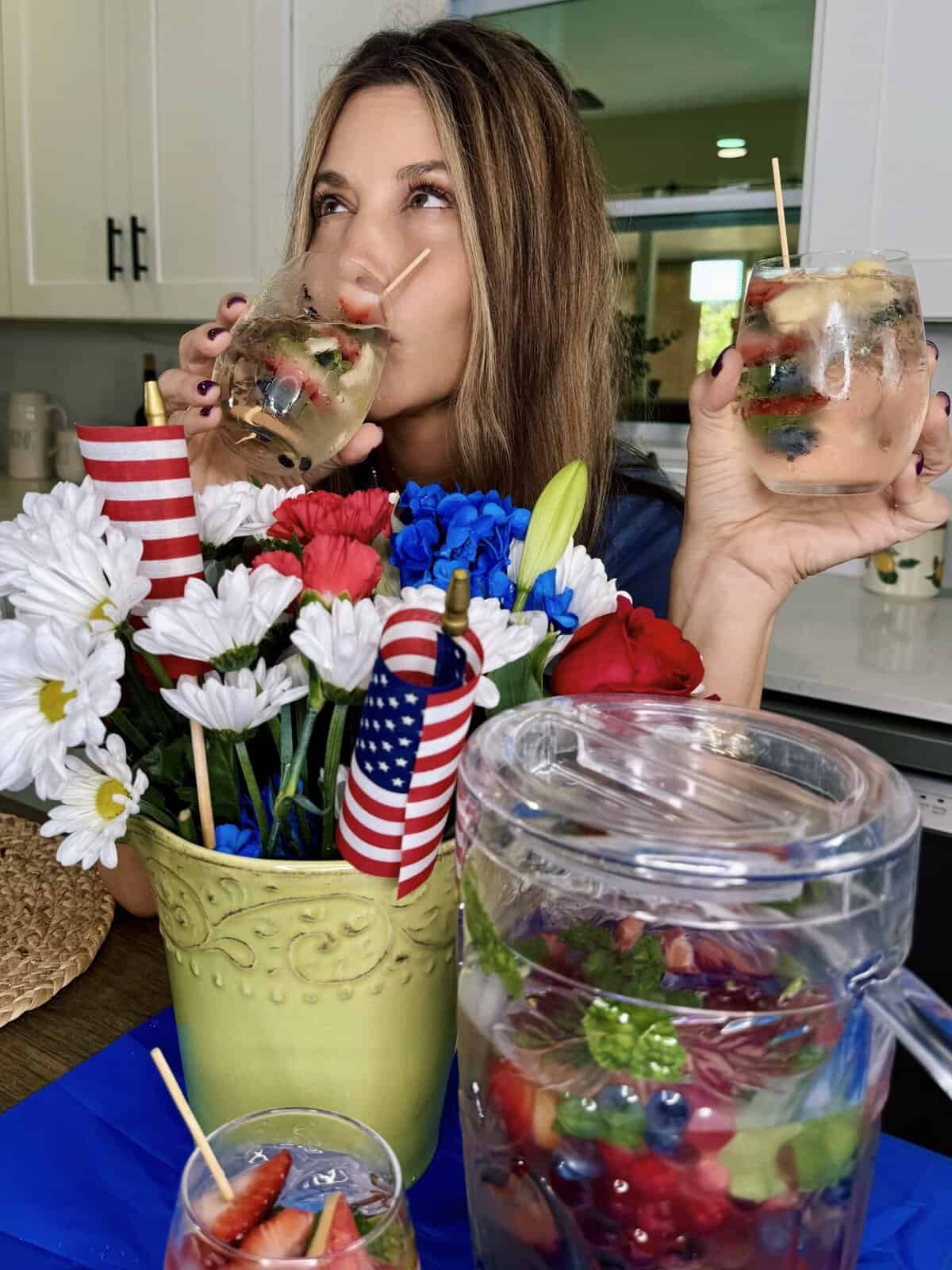 A woman holds two glasses of Red White and Boozy Pitcher Cocktail, sitting behind a green vase of red, white, and blue flowers with American flags. A pitcher of sangria with berries and mint sits on the table before her.
