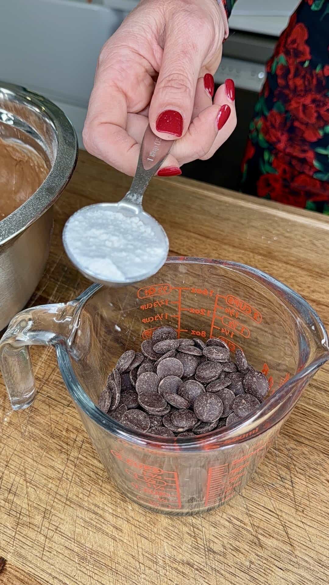 A hand with red nail polish holds a spoon of white powder above a measuring cup filled with chocolate chips on a wooden surface. A metal mixing bowl is partially visible to the left.