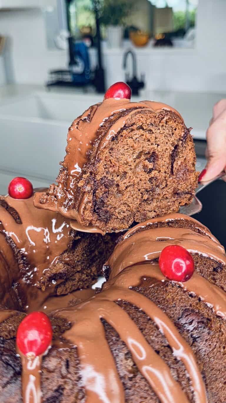 A close-up of a chocolate bundt cake topped with glossy chocolate icing and red berries. A hand is lifting a slice, revealing the moist, rich texture inside. A modern kitchen is blurred in the background.