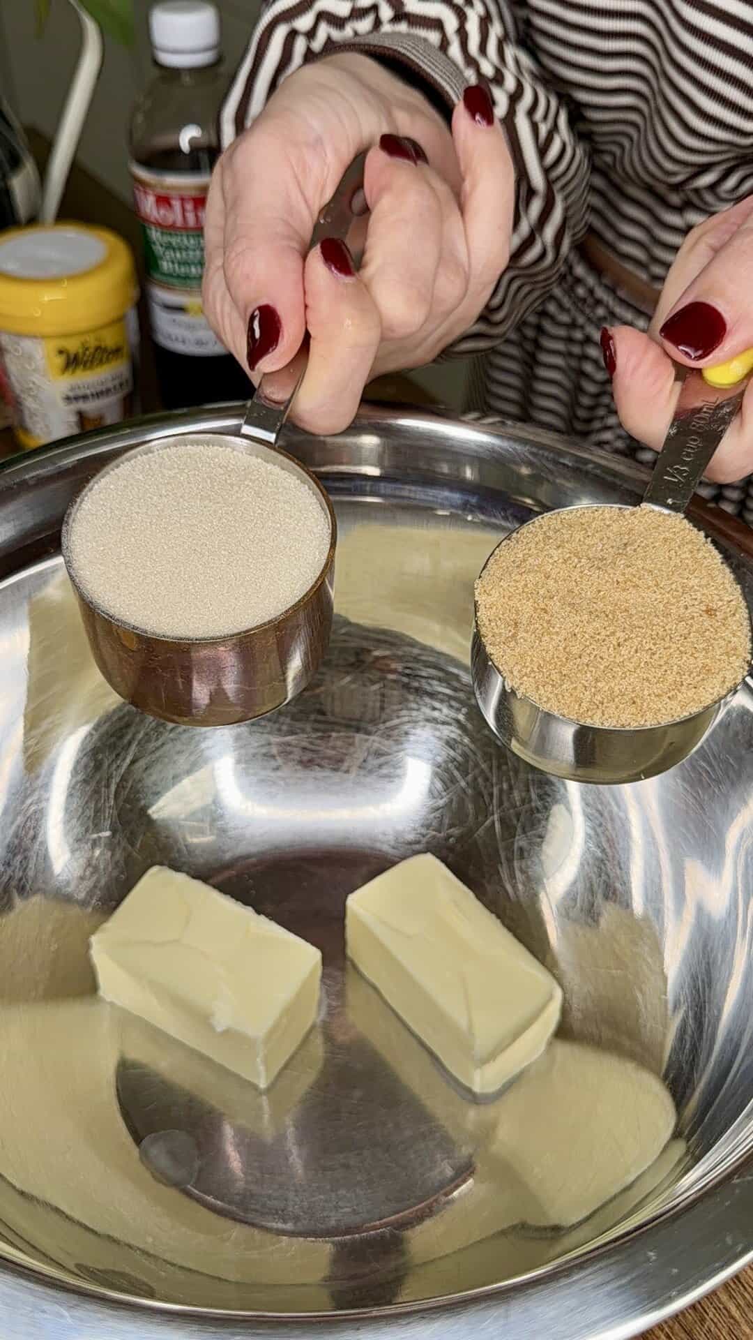 A person holds a measuring cup of granulated sugar and a measuring cup of brown sugar over a metal mixing bowl with two sticks of butter inside. Baking ingredients and containers are visible in the background.