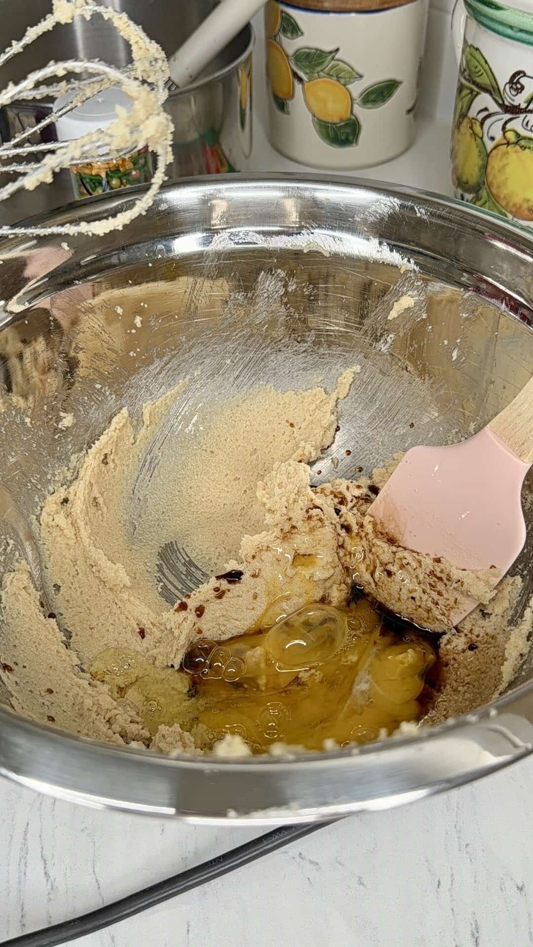 A metal mixing bowl with creamed butter and sugar, eggs, and vanilla being combined with a pink spatula; a hand mixer and kitchen containers with fruit designs are in the background.