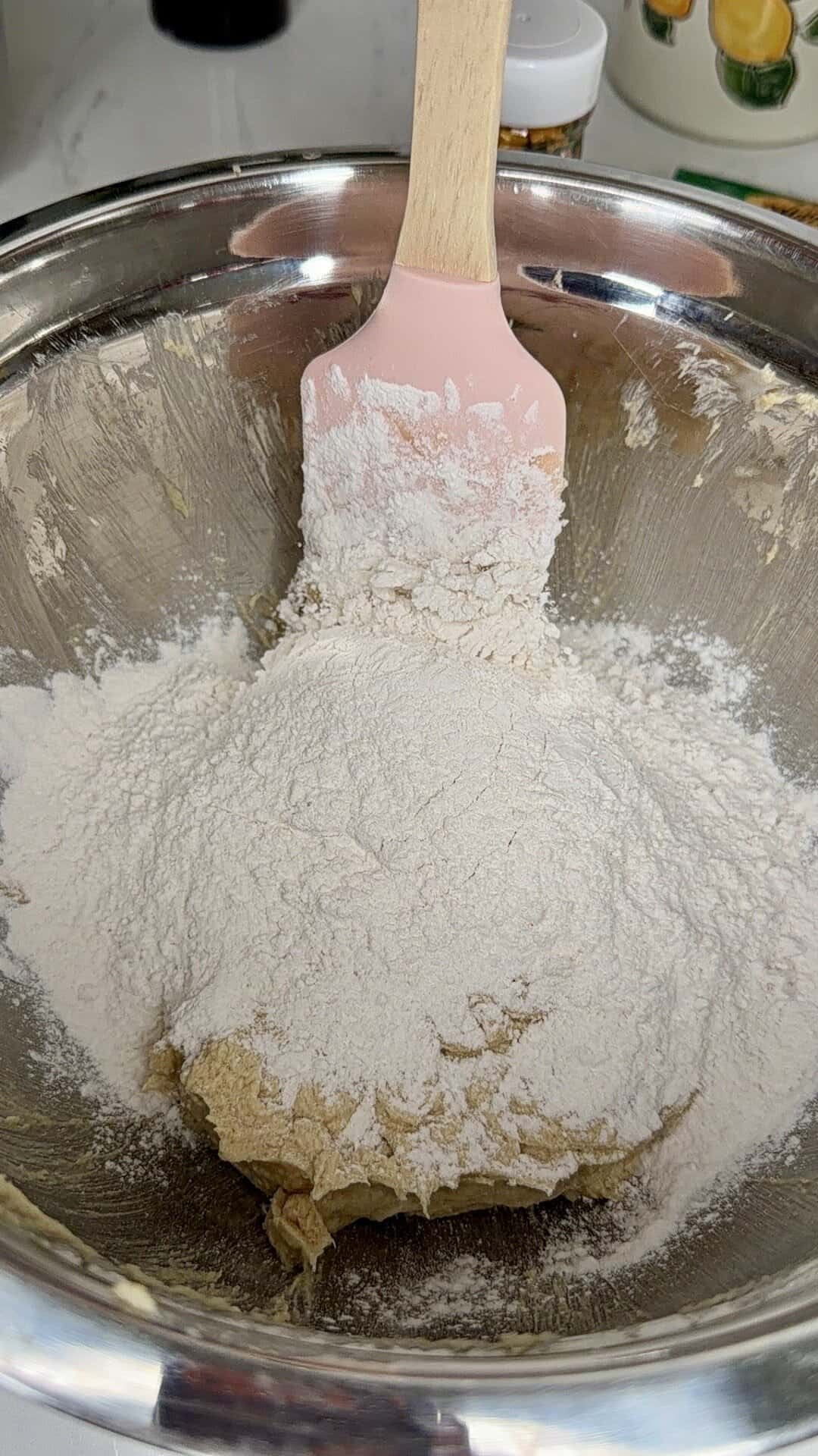 A pink spatula rests in a metal mixing bowl containing a mound of white flour on top of a brown dough mixture, ready to be combined for baking.