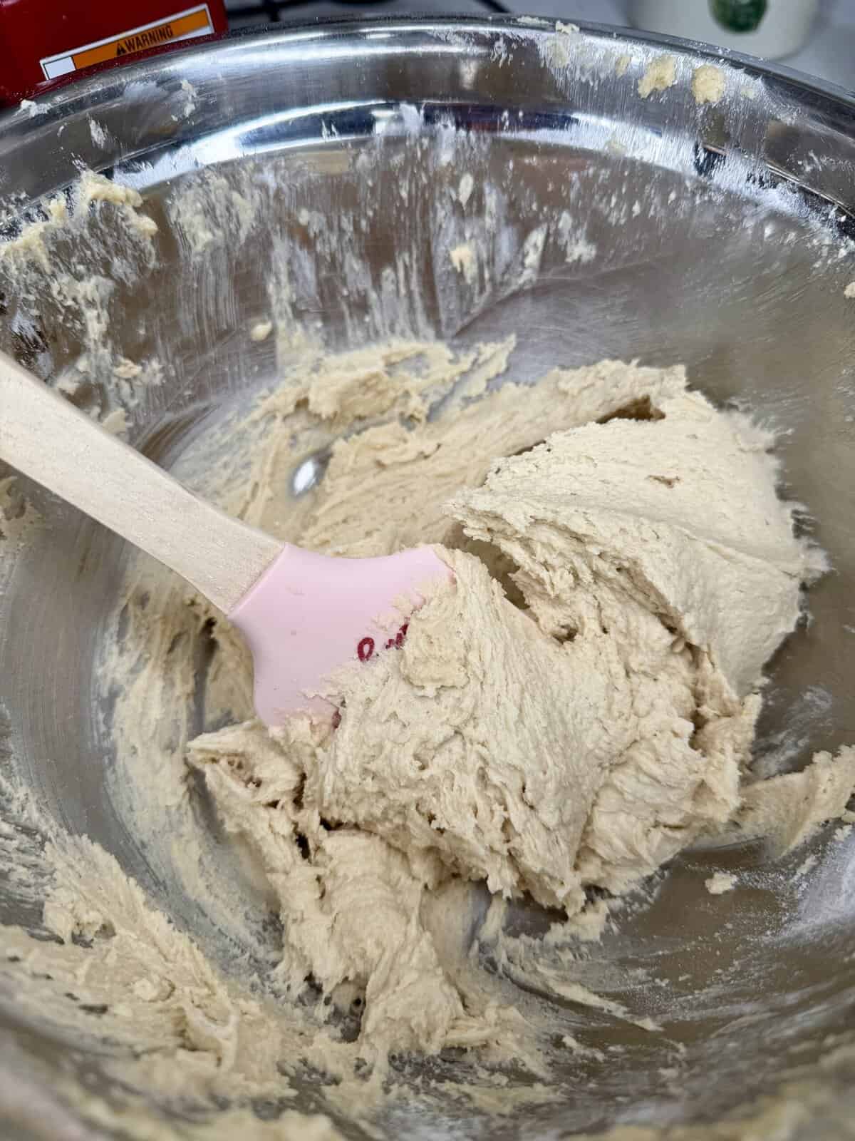 A pink spatula rests in a metal mixing bowl filled with light tan cookie dough, partially mixed and creamy in texture.