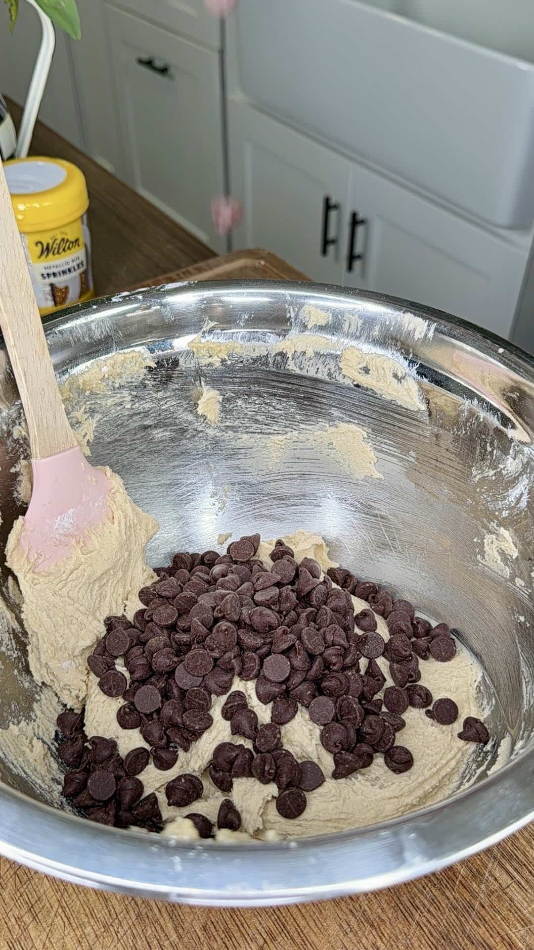 A large metal mixing bowl with cookie dough and chocolate chips, being stirred with a pink spatula on a wooden countertop in a kitchen. A yellow container is in the background near a white sink and cabinets.