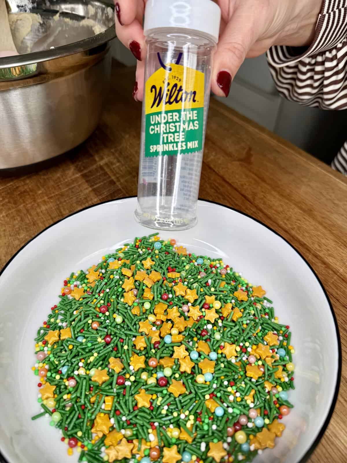A hand holds a Wilton Under the Christmas Tree sprinkles container above a bowl filled with colorful holiday sprinkles shaped like stars, beads, and rods. A metal mixing bowl is in the background on a wooden surface.