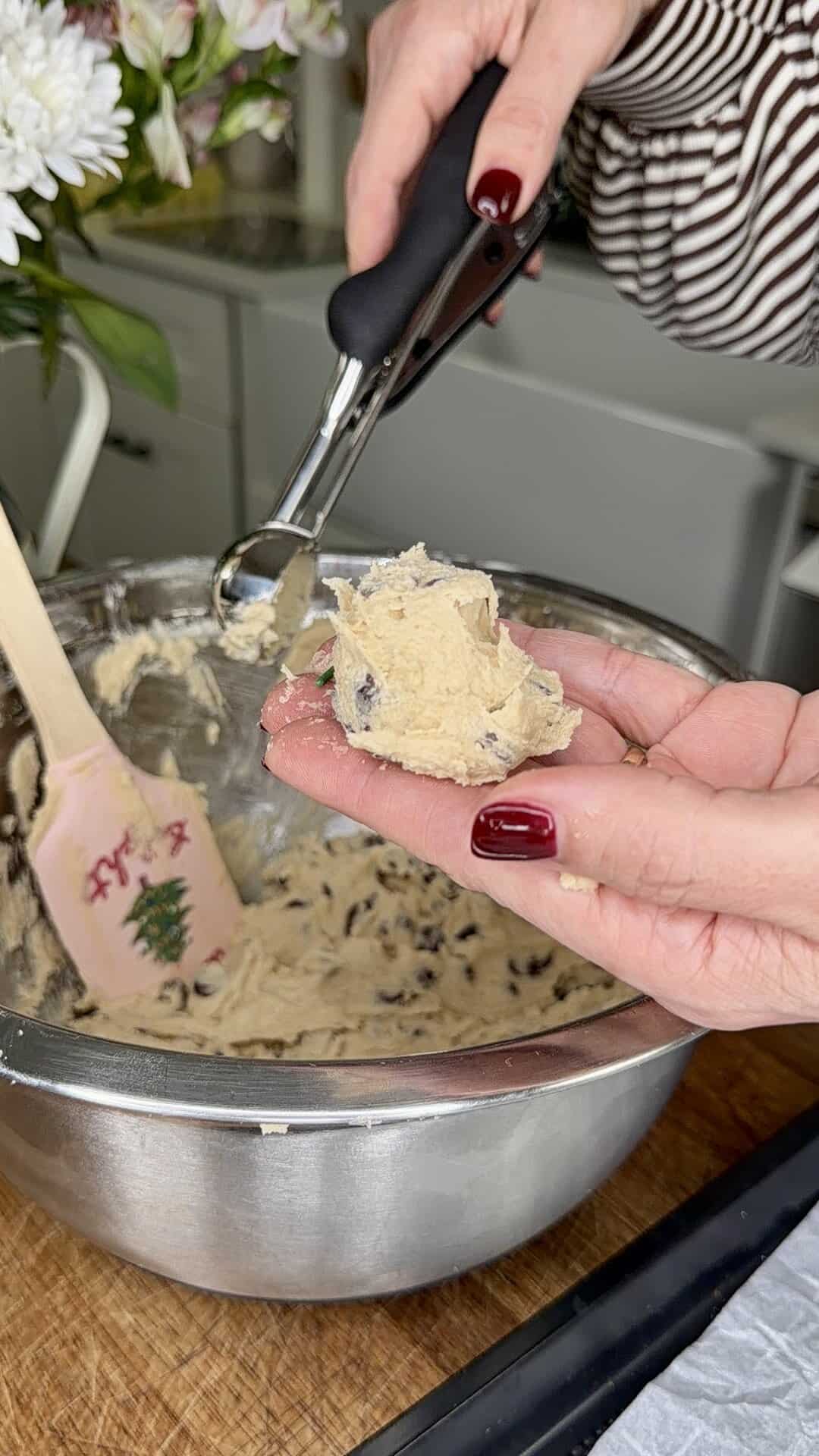 A person with red nail polish scoops raw chocolate chip cookie dough from a metal bowl using a cookie scoop. A festive spatula and flowers are visible in the background.