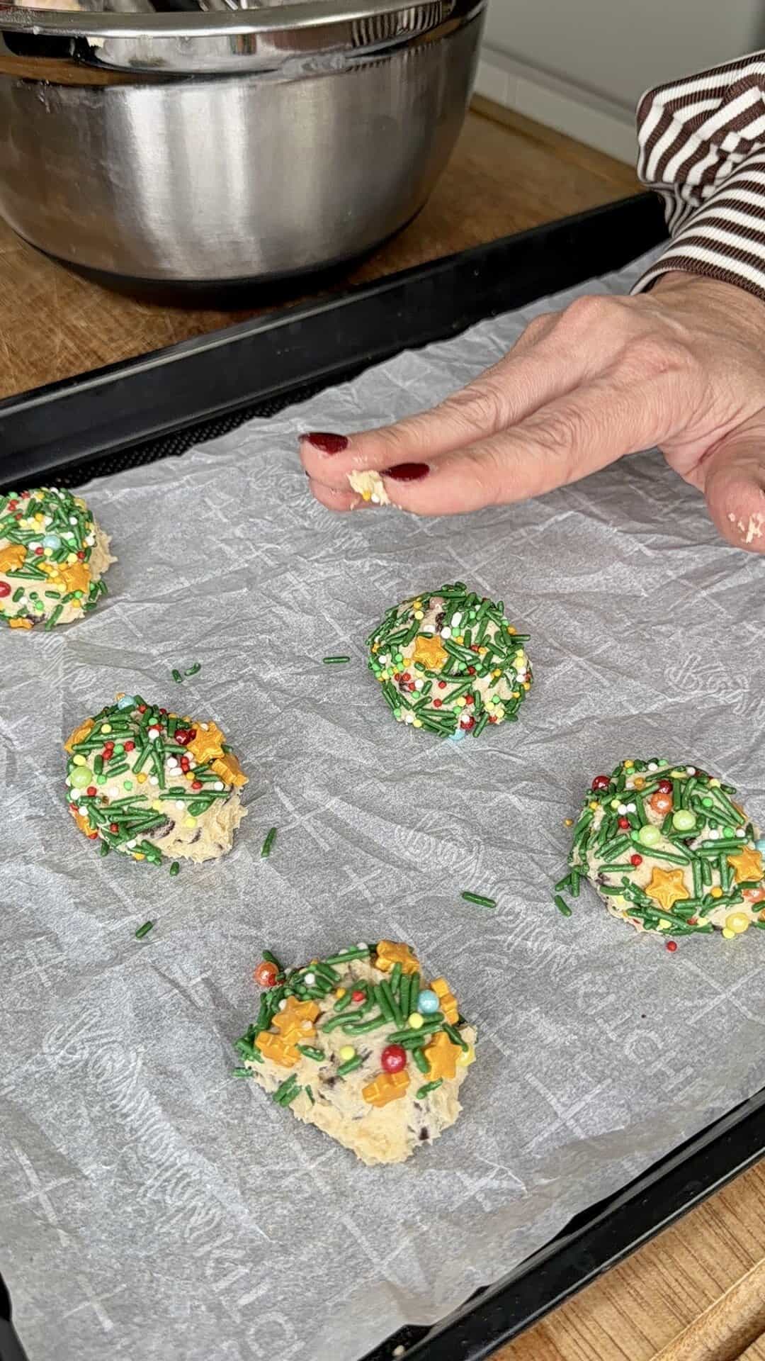 A hand places a ball of cookie dough covered in colorful sprinkles onto a parchment-lined baking tray. Several similar cookie dough balls are already on the tray, ready to be baked.