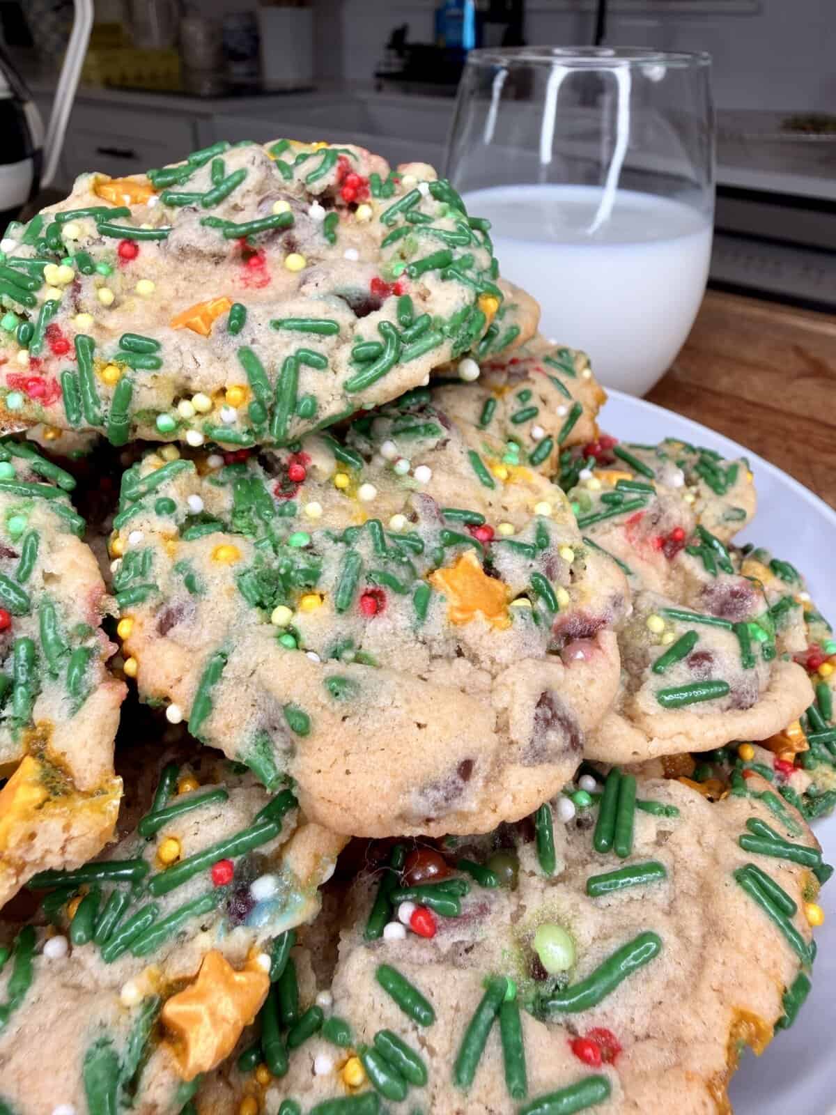 A plate of colorful sprinkle cookies with chocolate chips and festive decorations sits next to a glass of milk on a kitchen counter.