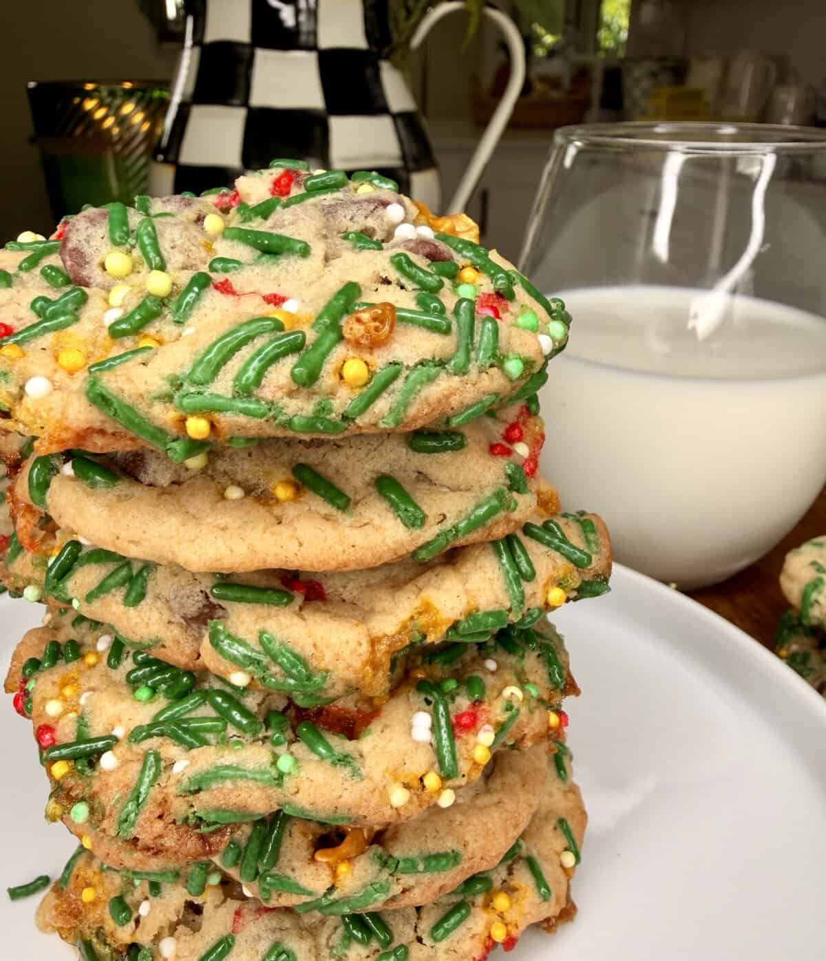 A stack of festive cookies with colorful sprinkles sits on a white plate next to a glass of milk, with a checkered teapot and kitchen background visible behind them.