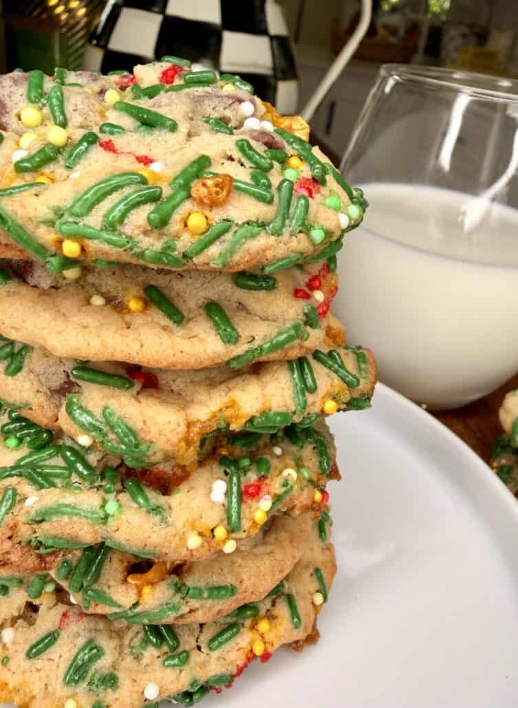 A stack of colorful sprinkle cookies on a white plate, with a glass of milk in the background. The cookies are decorated with green, yellow, white, and red sprinkles.