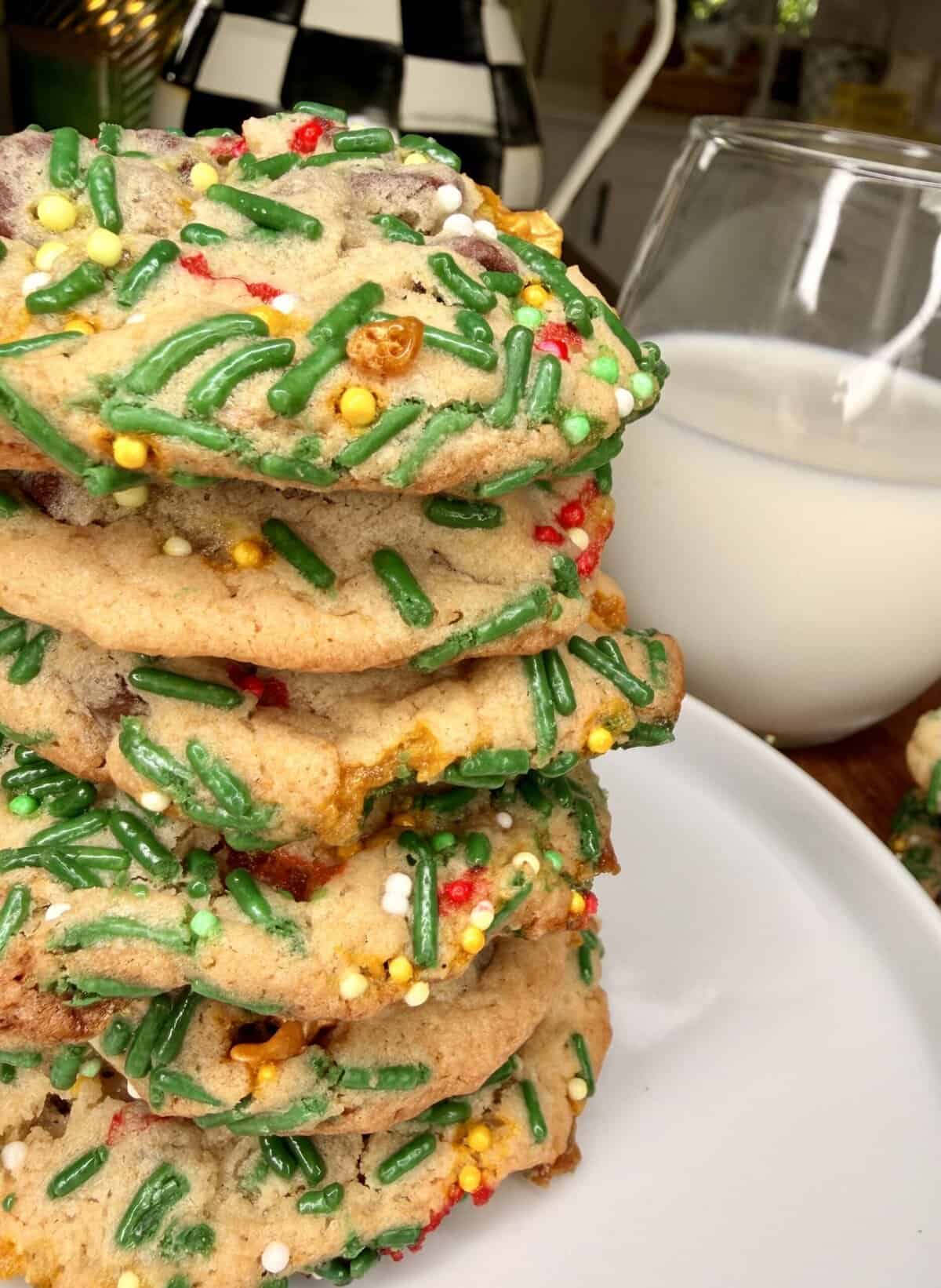A close-up of a stack of cookies covered with colorful sprinkles on a white plate, with a glass of milk in the background.