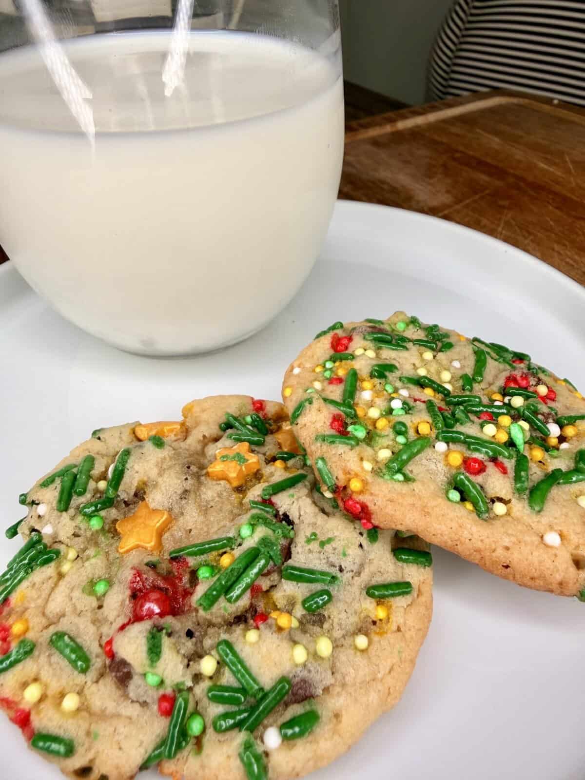 Two festive cookies with colorful holiday sprinkles sit on a white plate next to a glass of milk. The cookies have green, red, yellow, and white decorations, creating a cheerful, celebratory look.