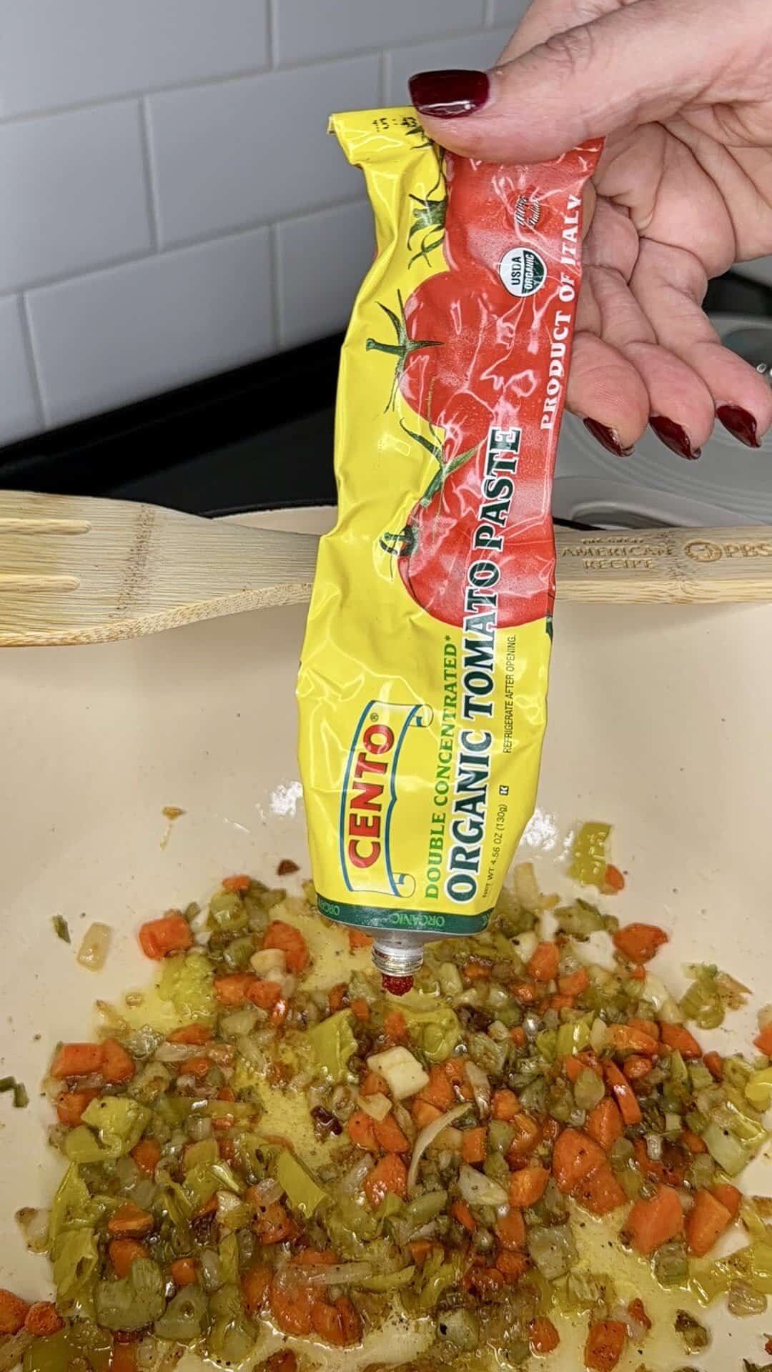 A hand with dark red nail polish squeezes a tube of Cento Organic Tomato Paste over a pan of sautéed chopped vegetables, including carrots, celery, onions, and peppers.