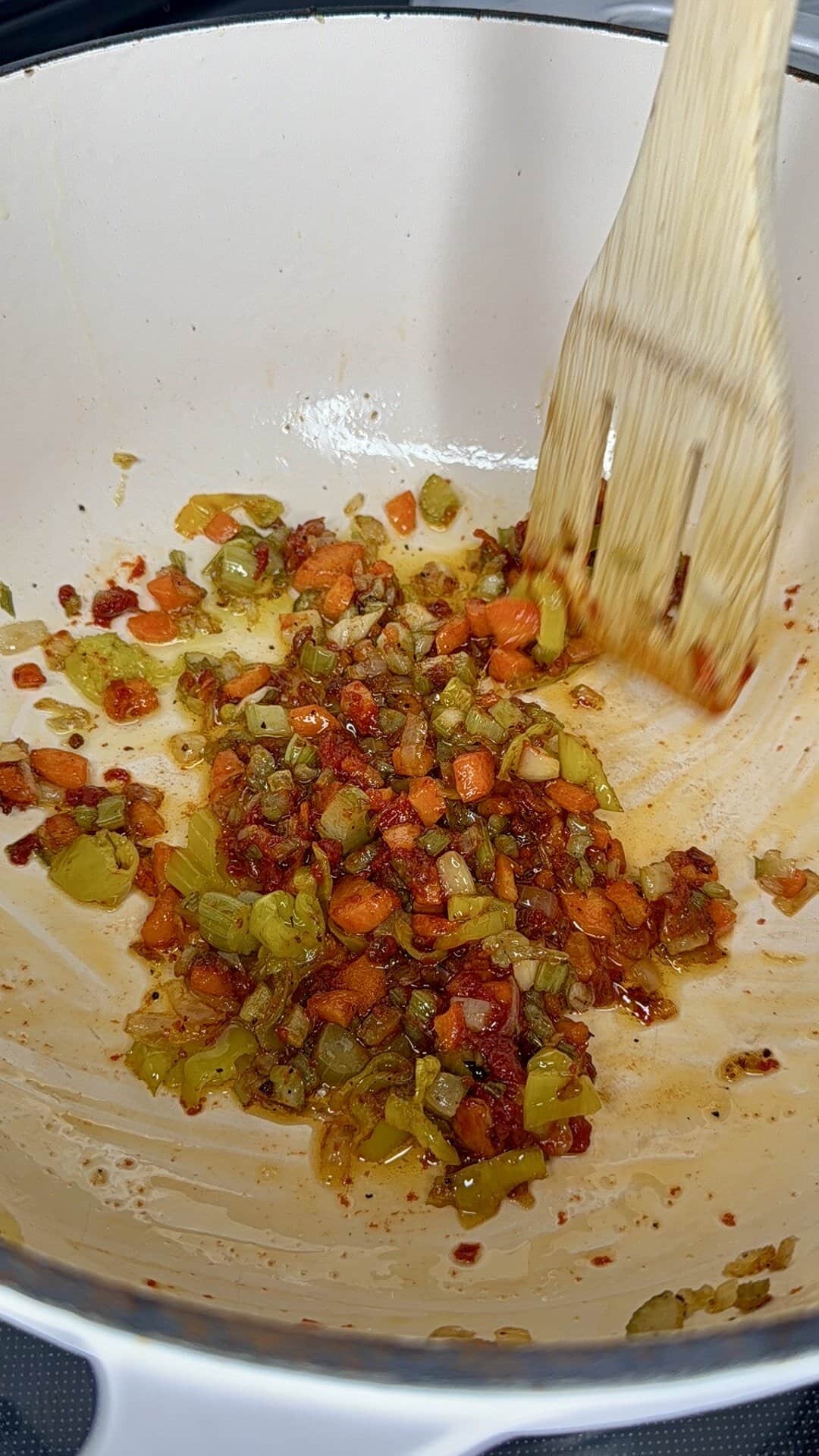 Diced vegetables, including carrots, celery, and bell peppers, are being sautéed in a white pot. A wooden spatula is stirring the mixture, which appears to be partially caramelized.