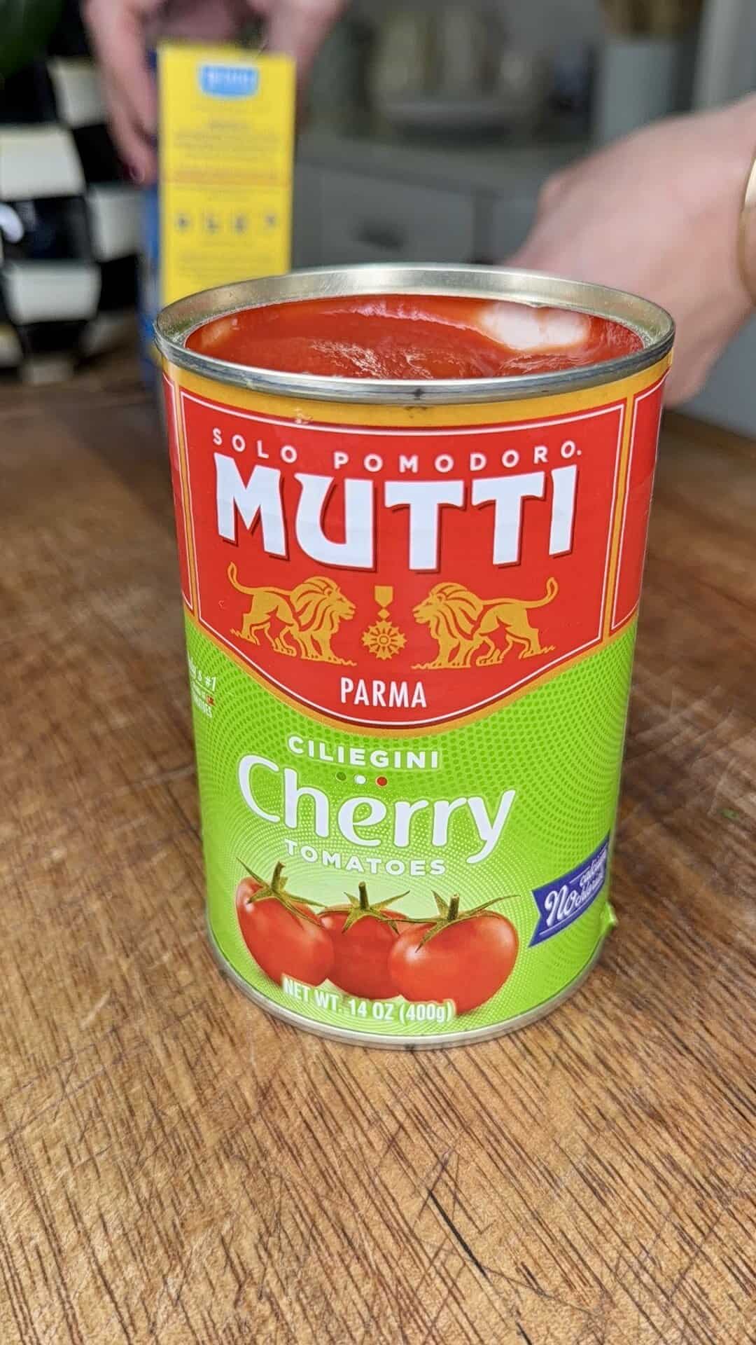 A close-up of an open can of Mutti Cherry Tomatoes sits on a wooden surface, with tomatoes and product details on the label. A person’s hand and a blurred box are visible in the background.