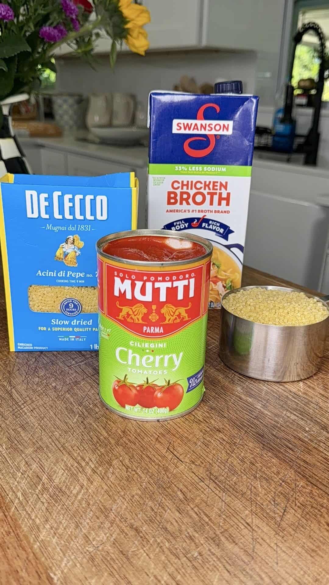 A box of De Cecco Acini di Pepe pasta, a can of Mutti cherry tomatoes, a carton of Swanson chicken broth, and a measuring cup filled with dry pasta on a wooden surface in a kitchen.