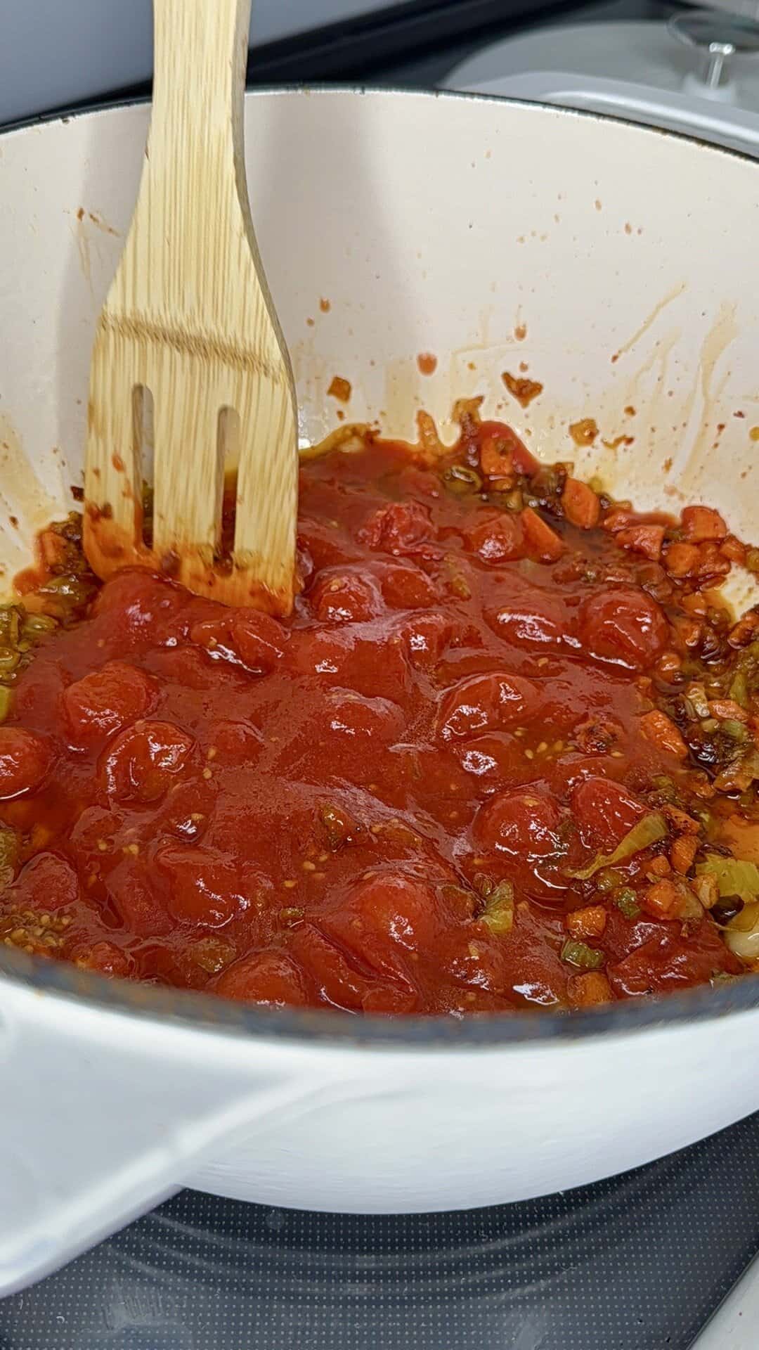 A wooden spatula stirs a pot of tomato sauce with chopped onions, carrots, and seasonings in a white Dutch oven on a stovetop.