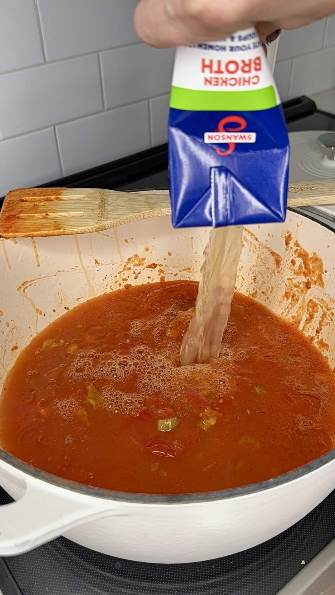 A hand pours chicken broth from a Swanson carton into a large white pot filled with tomato-based soup or sauce on a stovetop, with a wooden spoon resting on the pot’s edge.