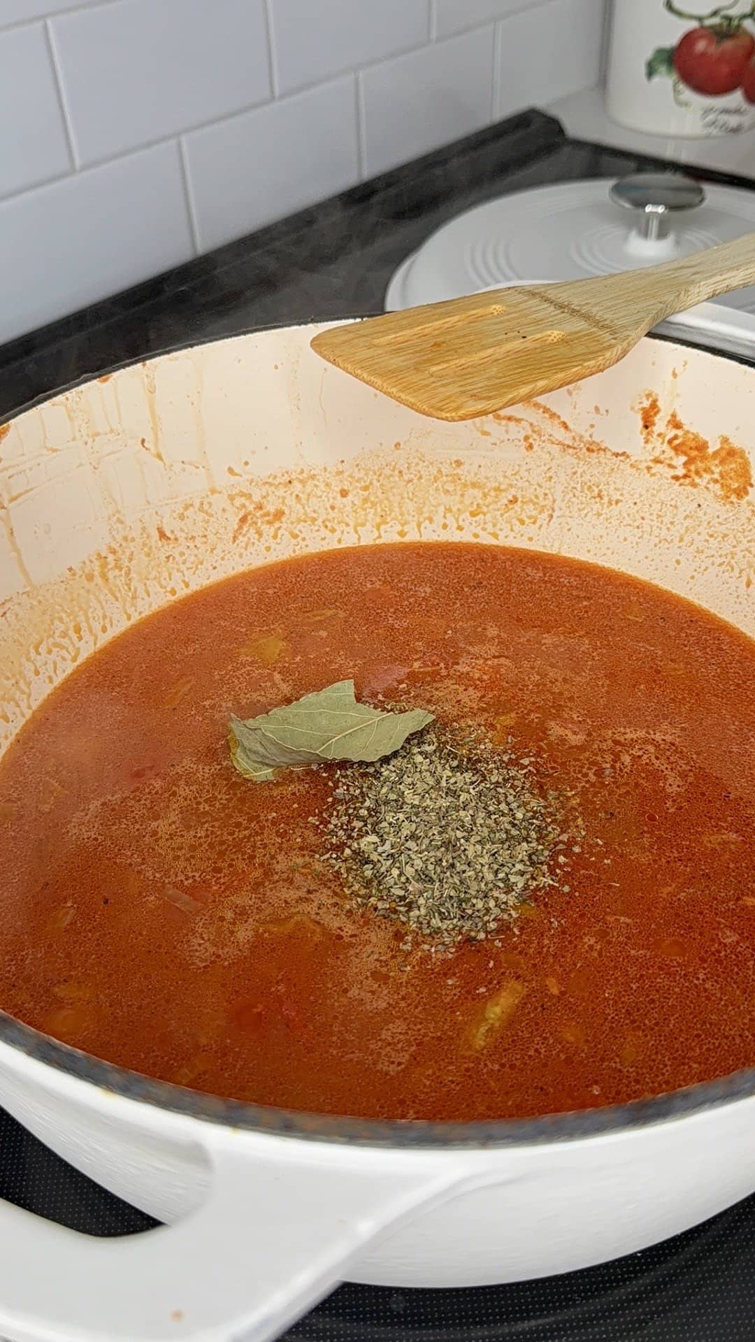 A white pot filled with simmering red tomato sauce on a stove, topped with dried basil and bay leaves; a wooden spoon rests on the side of the pot. Steam rises from the hot sauce.