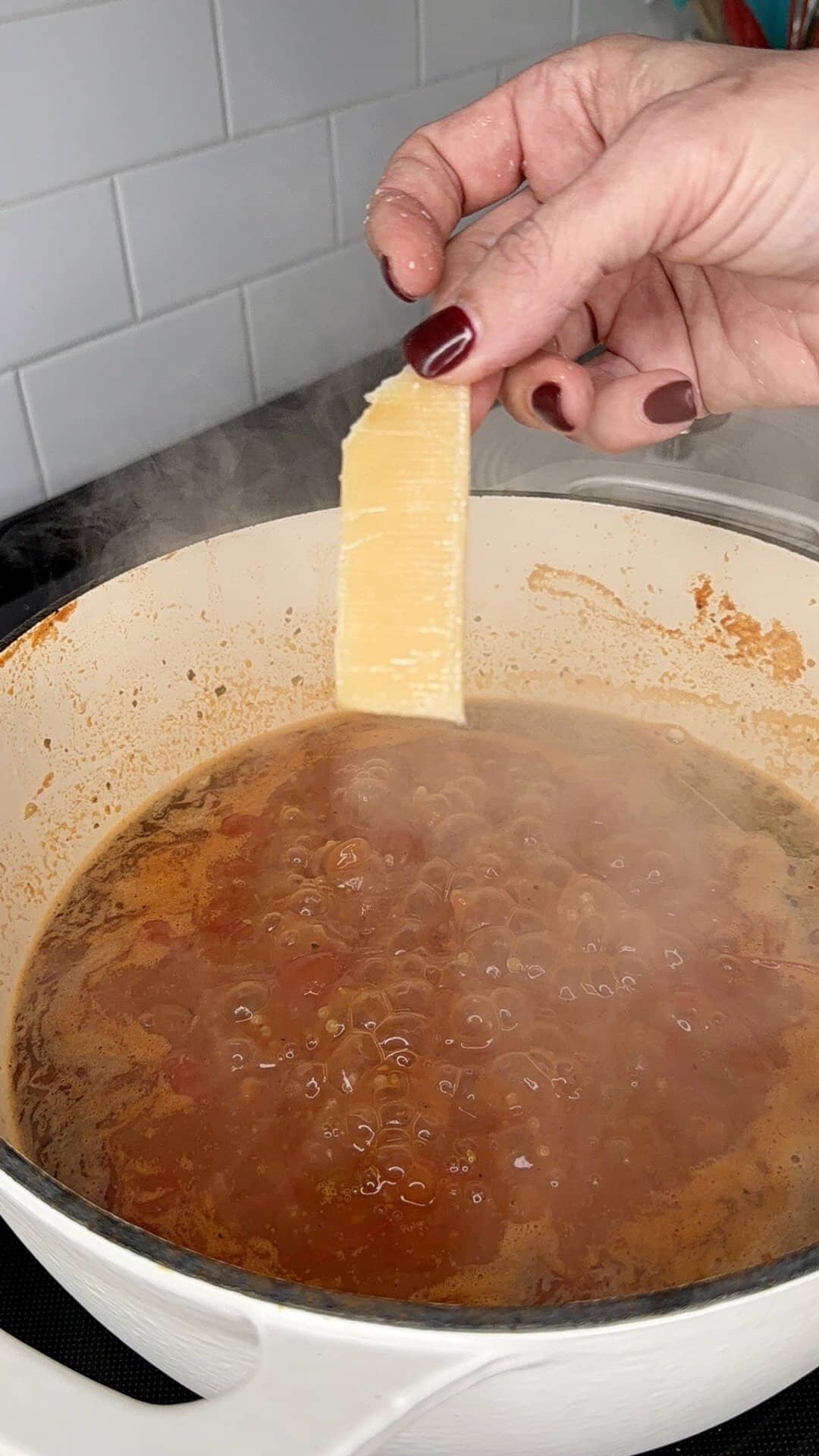 A hand with dark red nail polish holds a slice of Parmesan cheese over a steaming pot of tomato sauce simmering in a white Dutch oven on the stove.