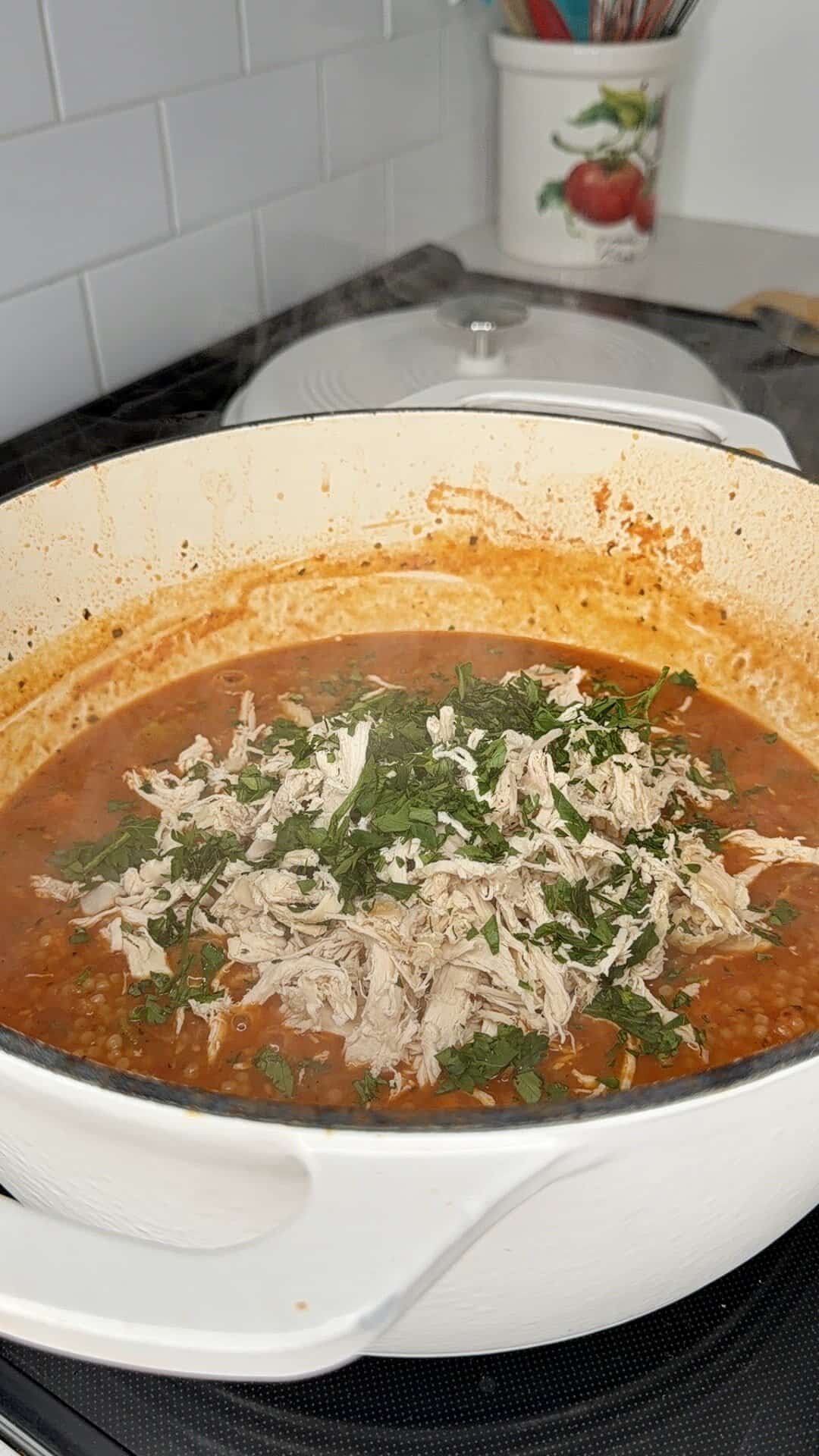 A white pot on a stove is filled with a red soup or stew, topped with shredded chicken and fresh chopped herbs. The lid rests in the background on a white countertop with a utensil holder nearby.