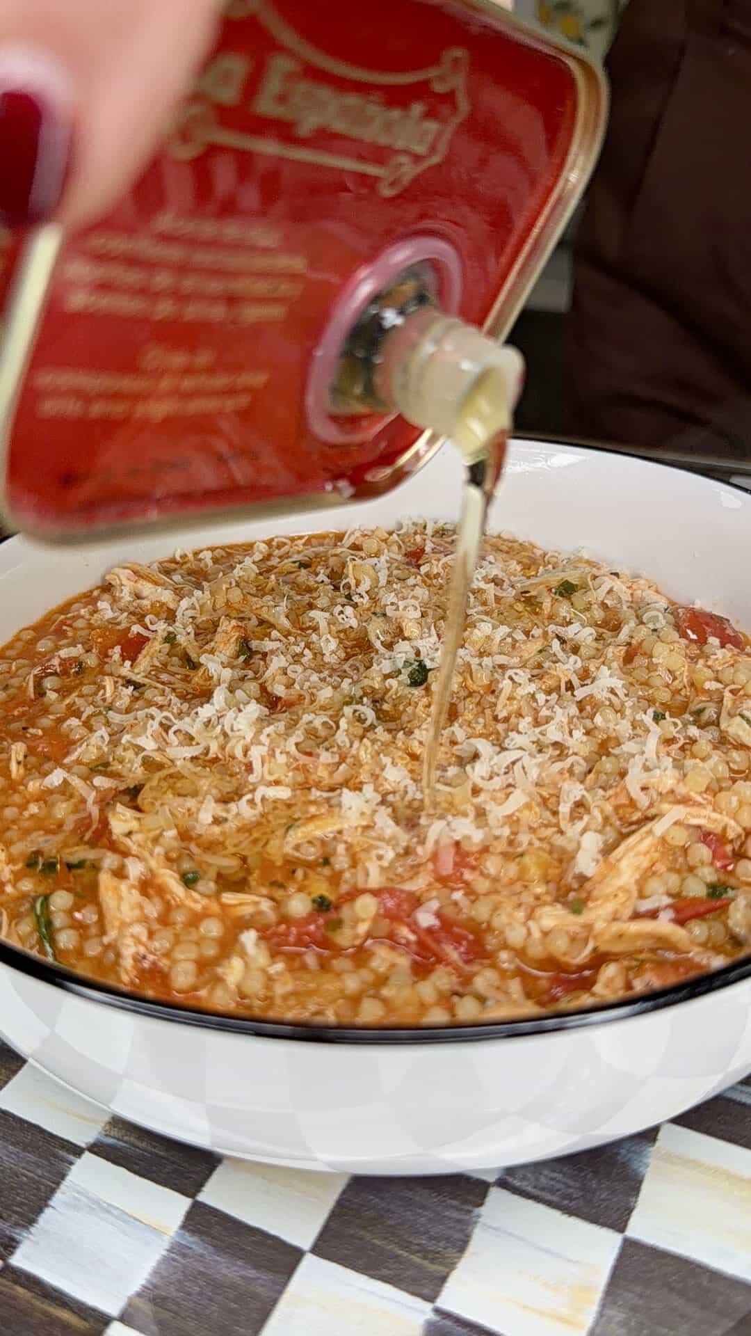 A hand pours olive oil from a red tin can onto a bowl of rice topped with grated cheese and tomato sauce, placed on a checkered surface.