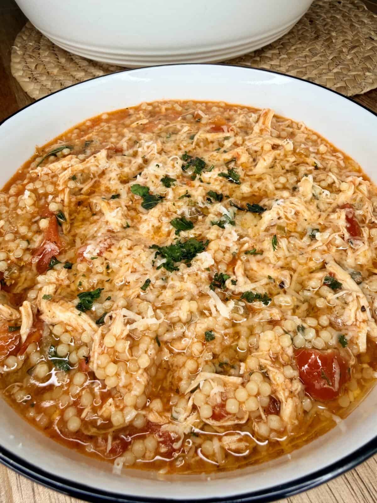 A bowl of chicken soup with pearl couscous, shredded chicken, tomatoes, and herbs, topped with grated cheese and garnished with chopped parsley.