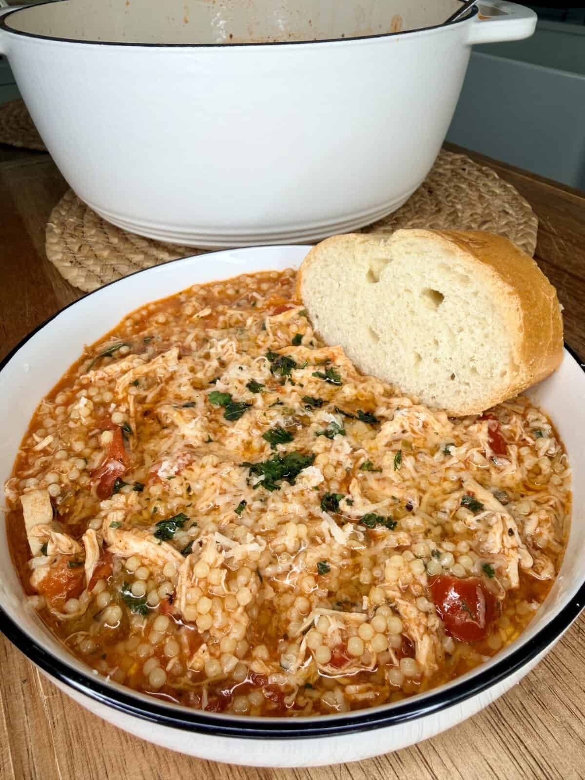 A bowl of tomato-based soup with shredded chicken, couscous, and vegetables, garnished with herbs, served with a slice of bread on the rim; a white pot is in the background.