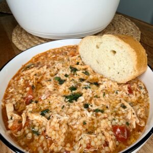 A bowl of tomato-based soup with shredded chicken, pearl couscous, and herbs, served with a slice of crusty bread. A white pot sits in the background on a woven mat.