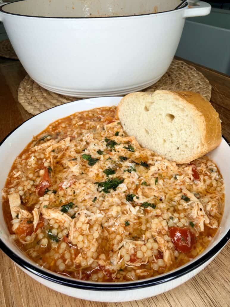 A bowl of tomato-based soup with shredded chicken, pearl couscous, and herbs, served with a slice of crusty bread. A white pot sits in the background on a woven mat.