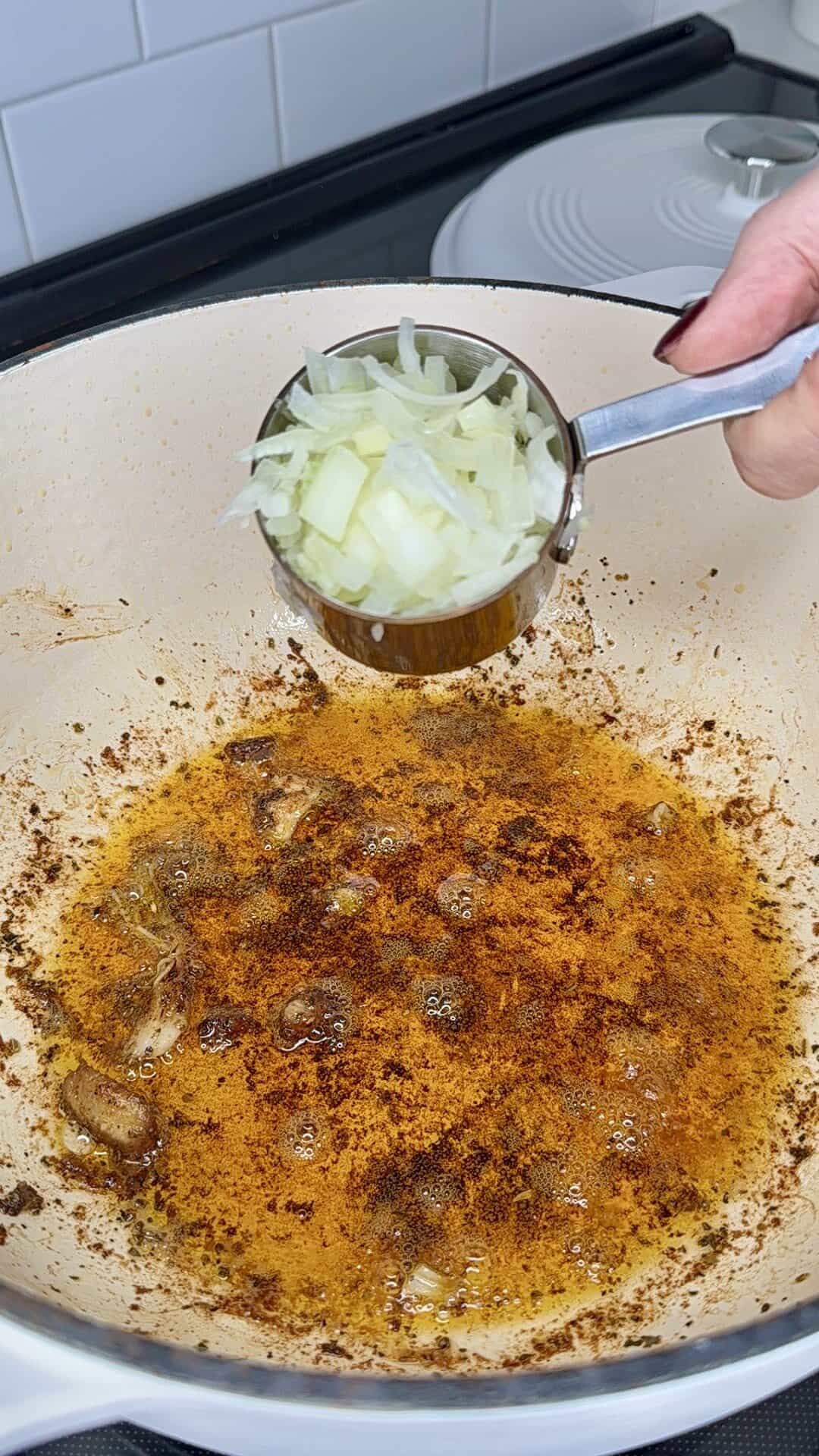 A hand holds a measuring cup filled with chopped onions above a pot with sizzling oil and browned bits on the bottom, ready for sautéing.