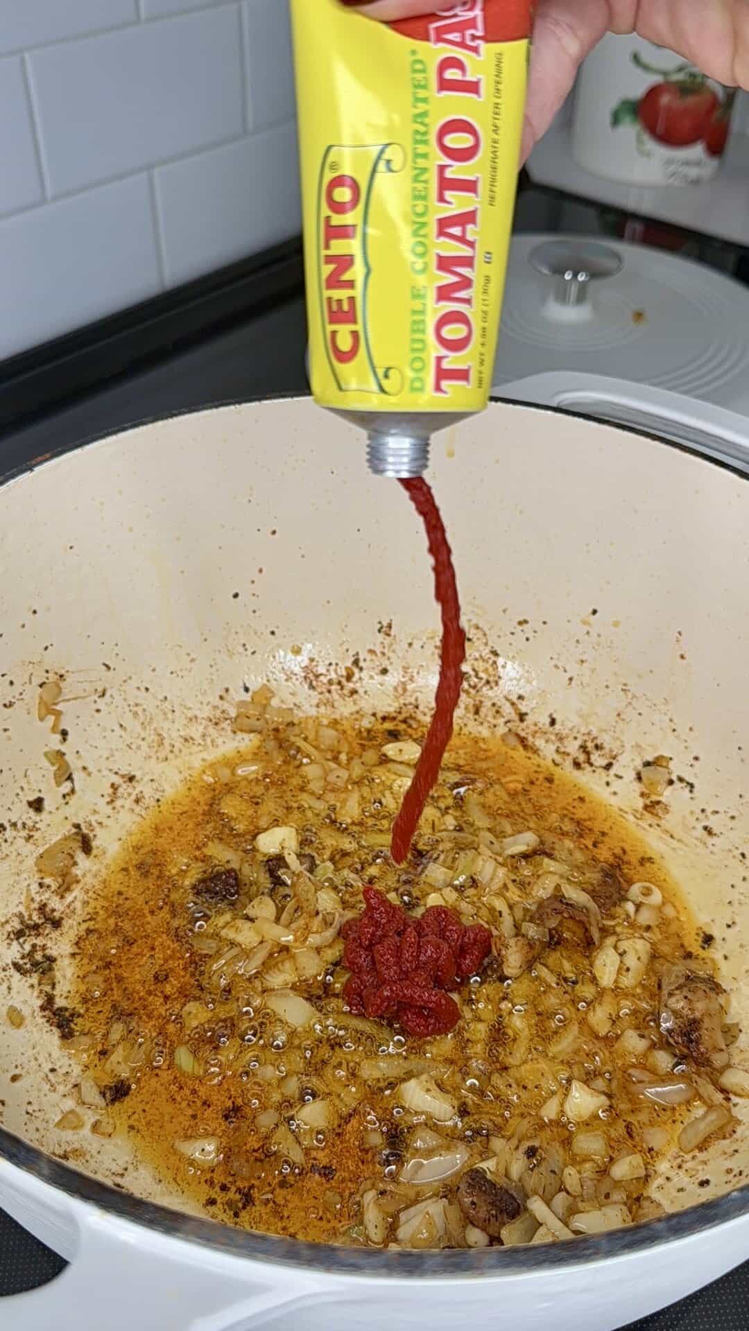 A hand squeezes Cento double concentrated tomato paste from a tube into a white pot containing sautéed onions and oil on a stovetop.