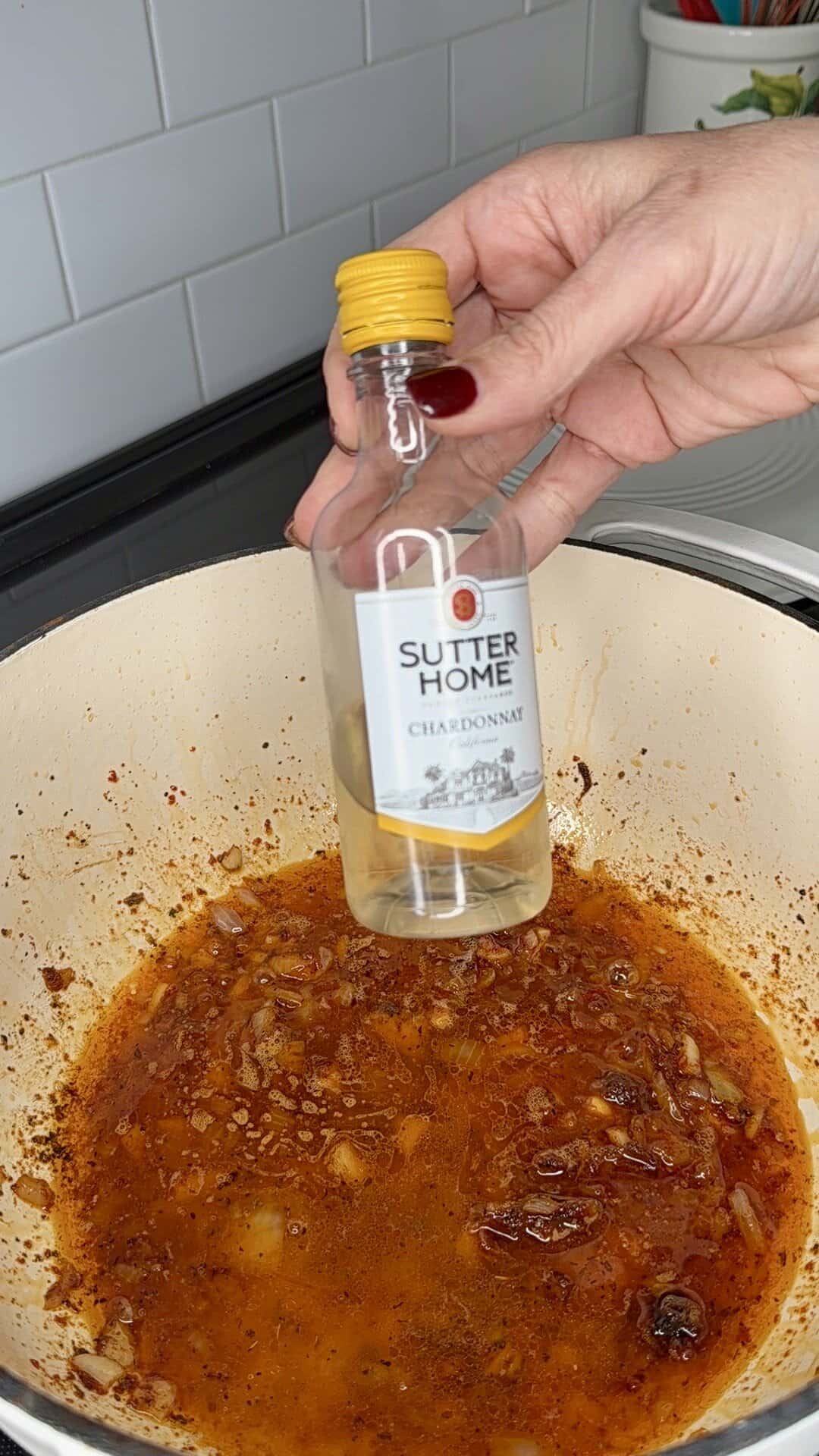 A hand holds an empty Sutter Home Chardonnay wine bottle over a white pot with sautéed onions and browned bits on the stovetop.