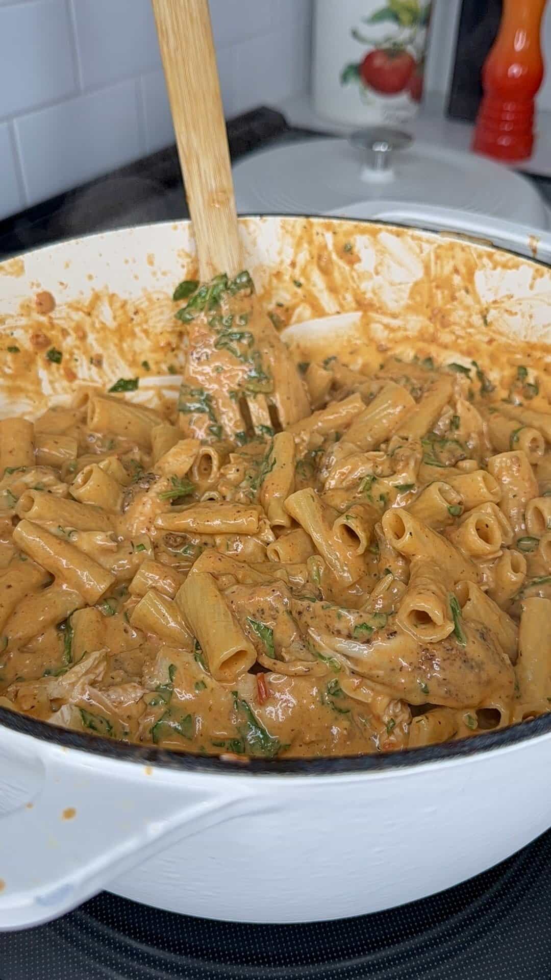 A creamy tomato pasta with penne, shredded chicken, and spinach being stirred with a wooden spoon in a large white pot on a stovetop.