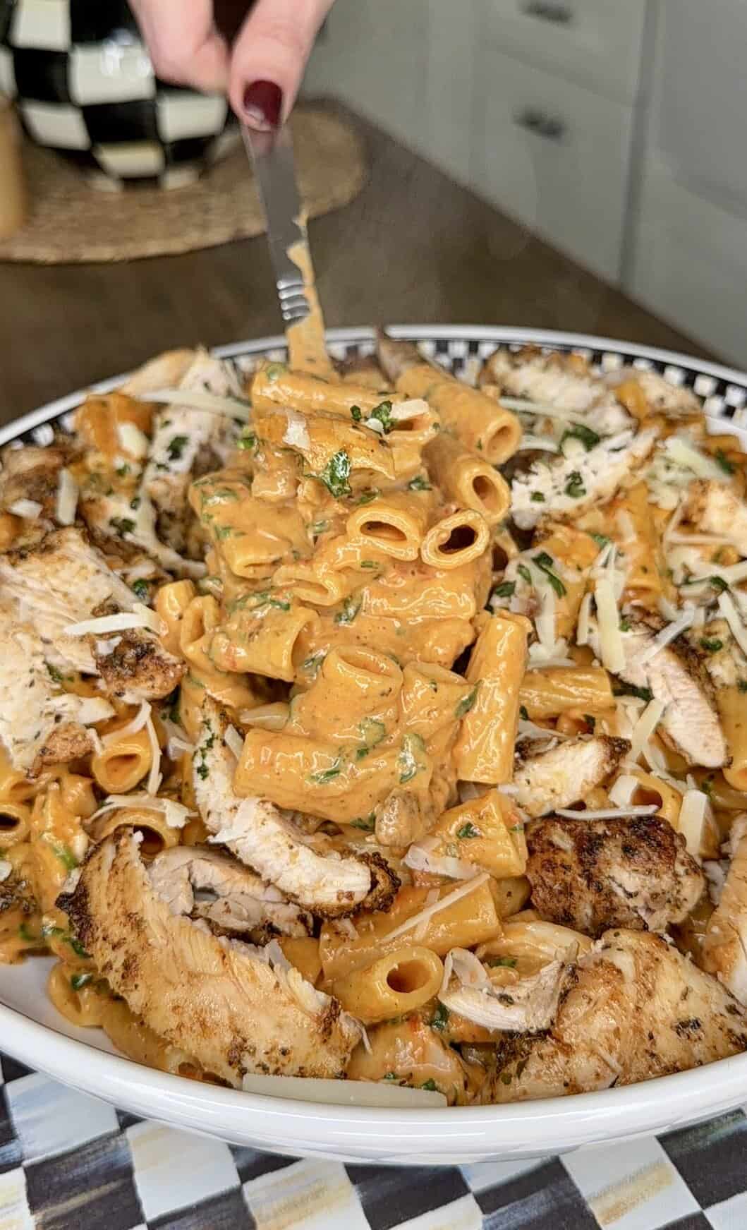 A close-up of creamy pasta with grilled chicken slices, garnished with herbs and shredded cheese. A hand holding a fork lifts a serving from a decorative bowl in a kitchen setting.